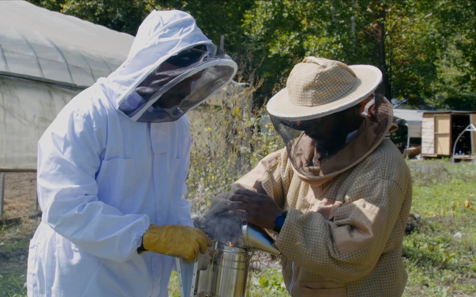 Two beekeepers in protective suits lighting a smoker beside a high tunnel at the DRT.