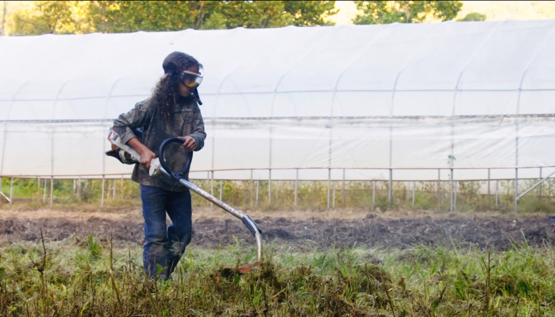 A young person in safety goggles using a string trimmer in front of a high tunnel.