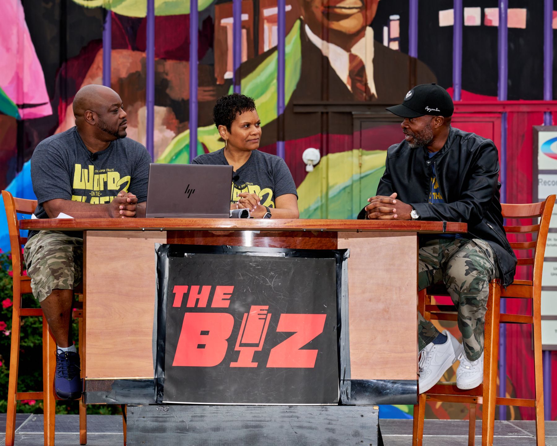 Jason Tartt, Sr. speaking with two hosts at a desk on an outdoor stage in front of a colorful mural.