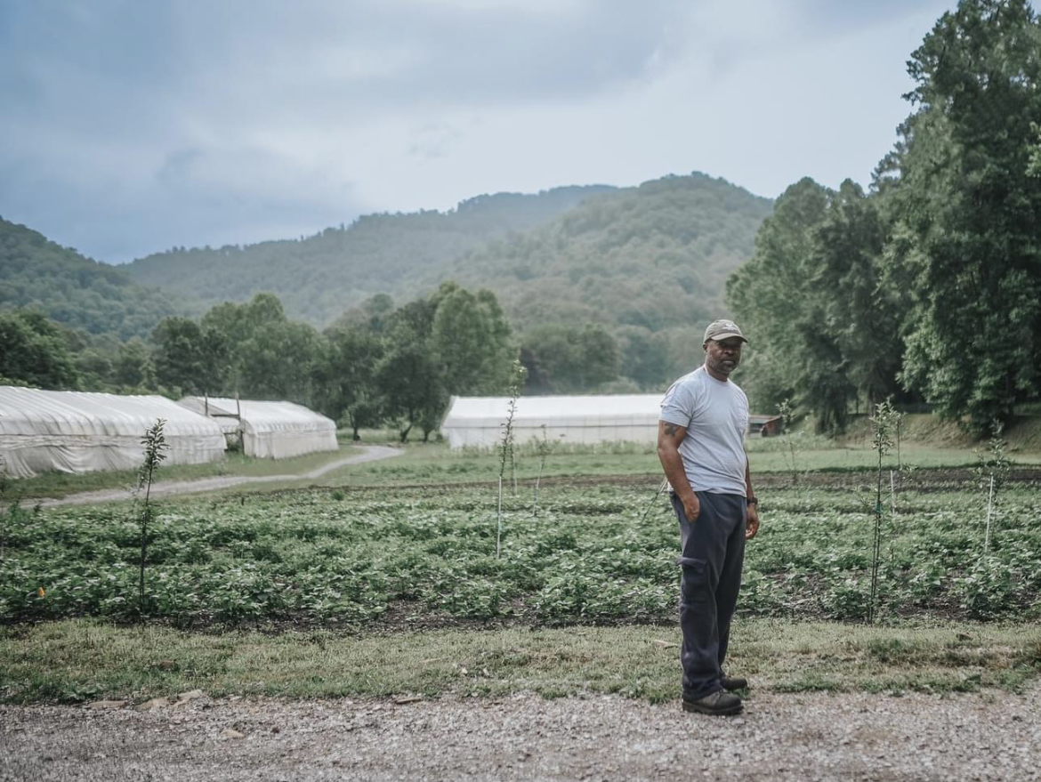 A farmer beside growing beds and high tunnels with wooded mountains behind.